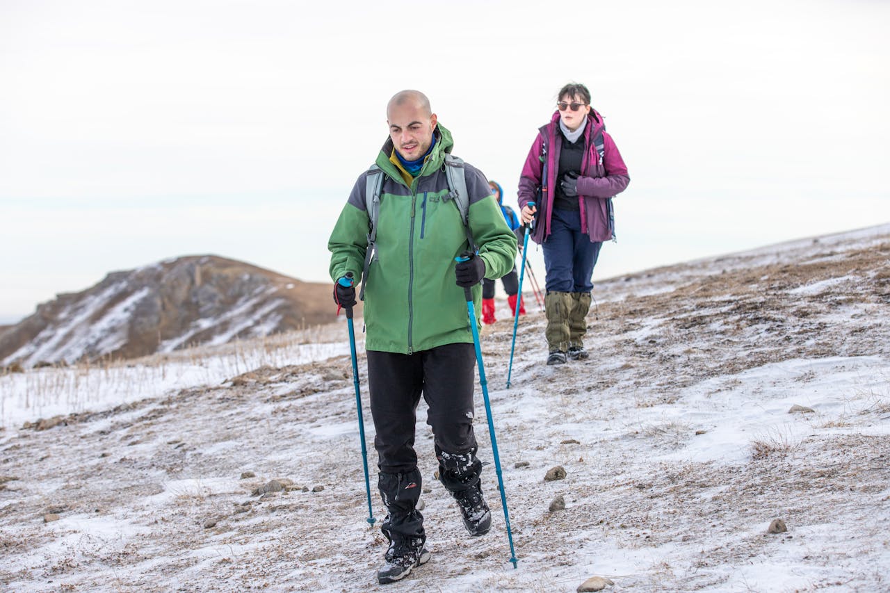 Two hikers trekking through a snowy hillside with hiking poles and winter gear.